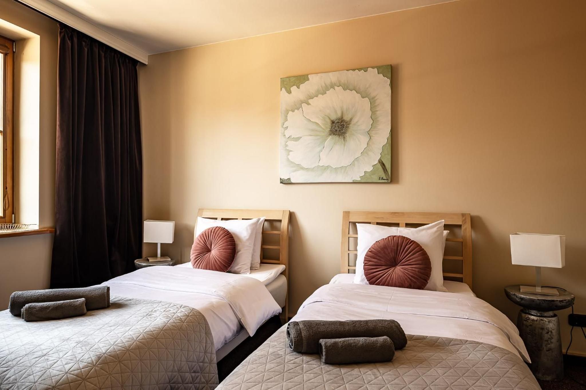 Ein Hotelzimmer mit zwei Betten, jeweils mit Kopfkissen und Decke, einem Nachttisch mit Lampe sowie einem großen Blumenbild an der Wand.