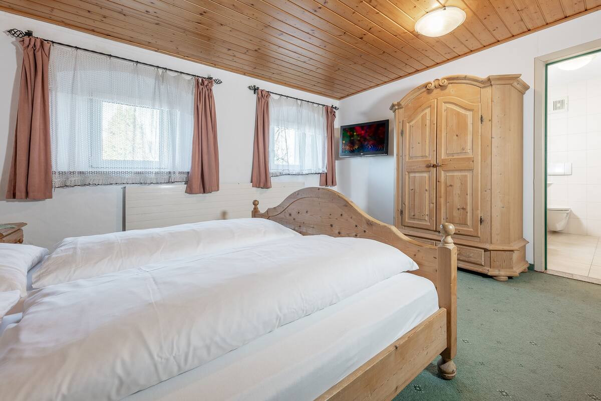Un dormitorio con cama de madera, dos ventanas con cortinas, un armario y un televisor en la pared.