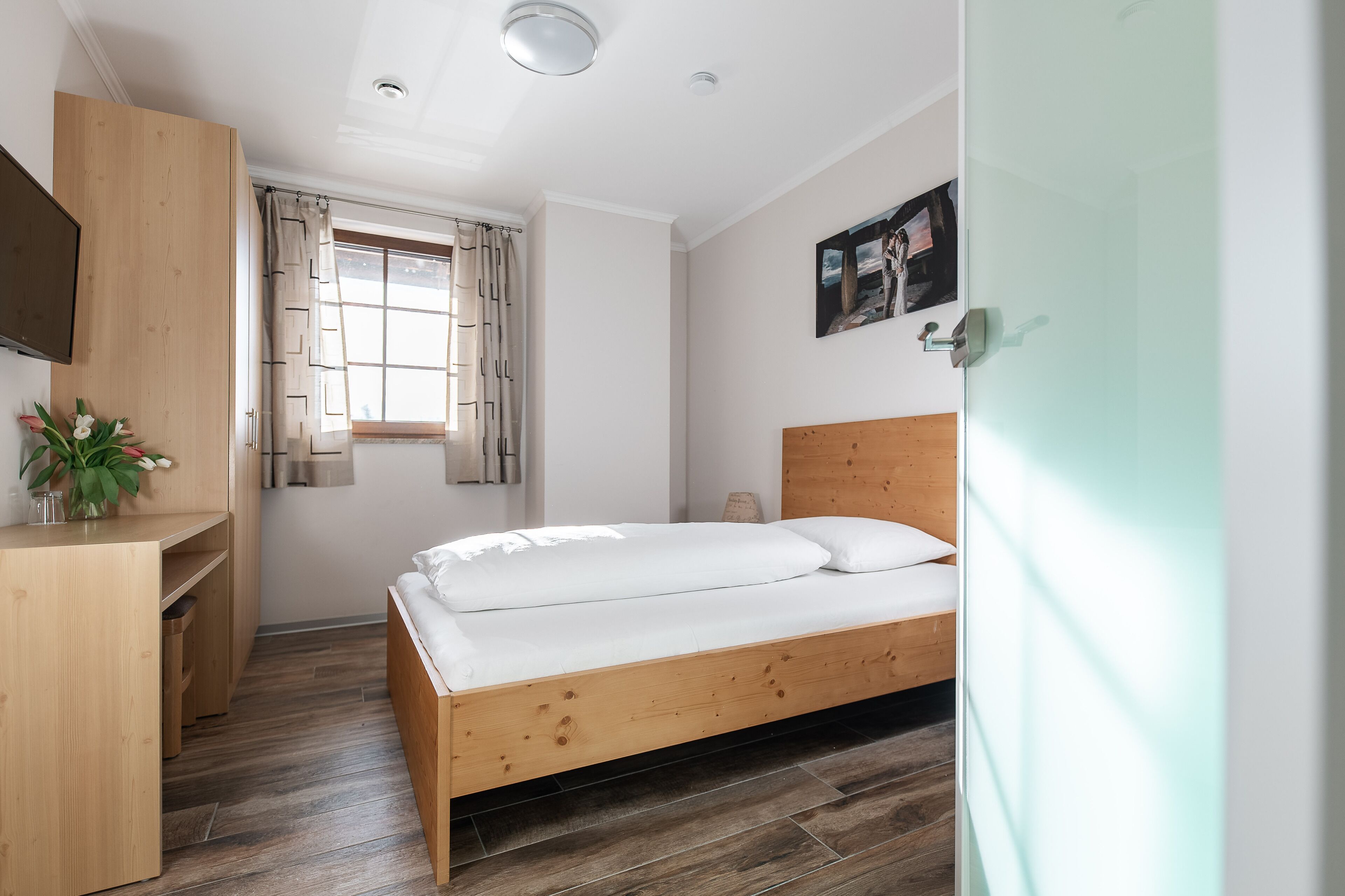 Une chambre à coucher avec un lit en bois, un bureau, une chaise, une fenêtre avec des rideaux et un téléviseur fixé au mur.