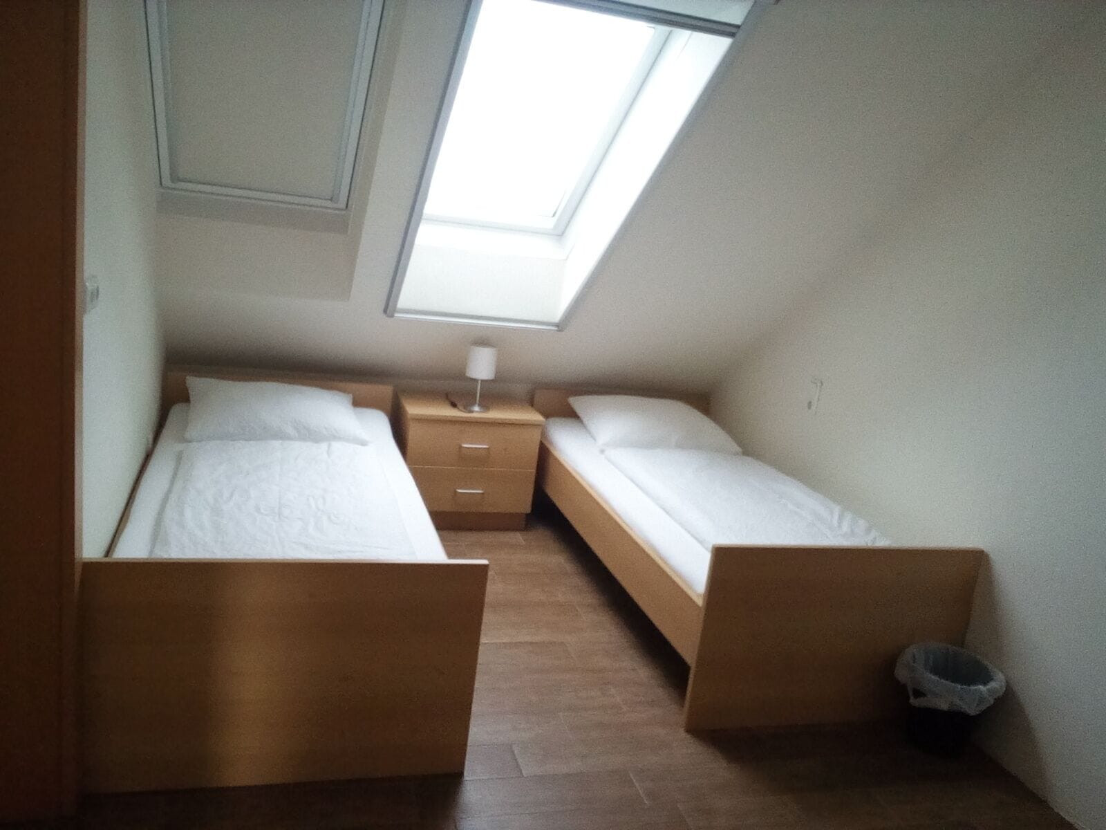 Una habitación pequeña y acogedora con dos camas individuales, una mesita de noche y un ventanal que proporciona luz natural.