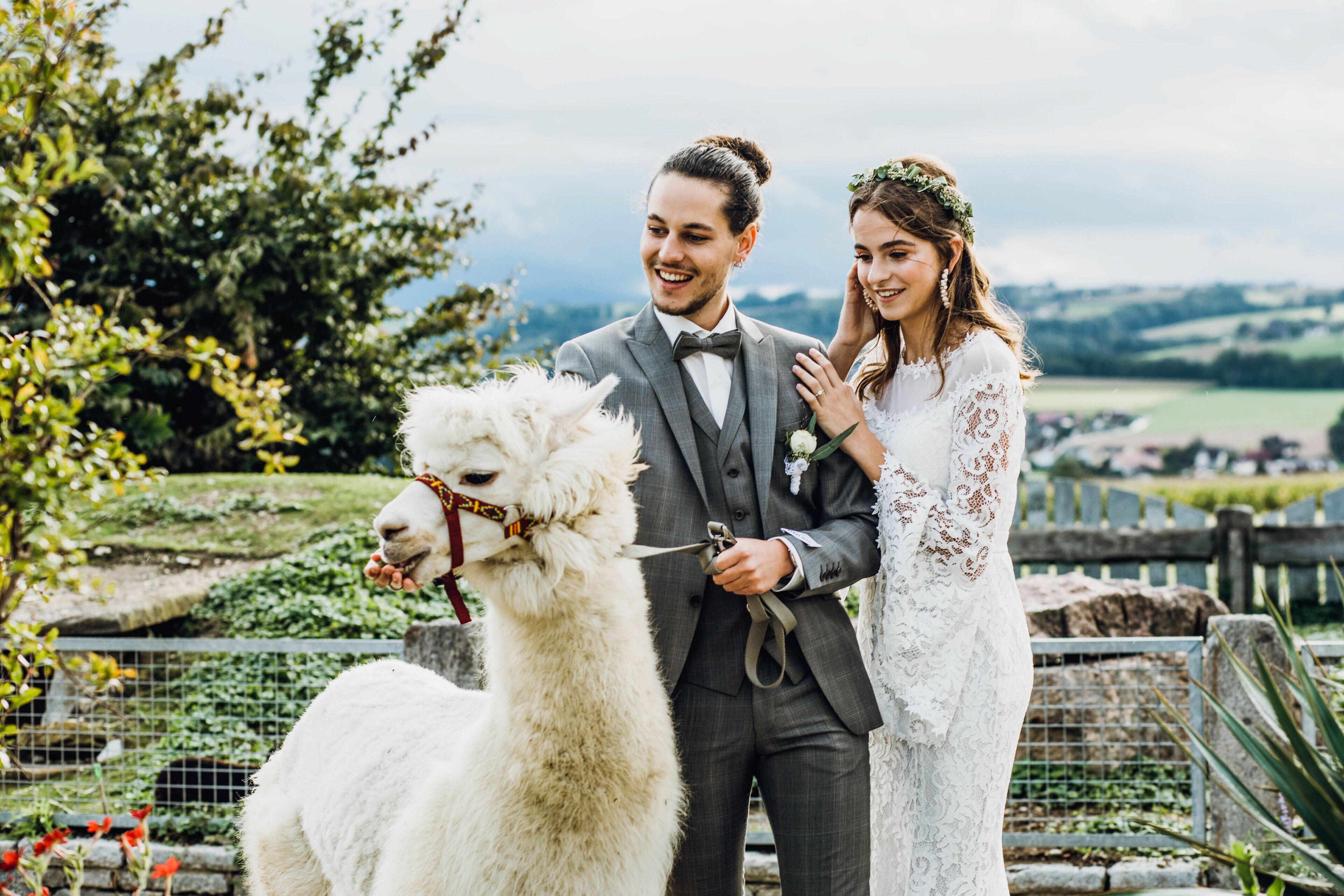 Una pareja vestida de novios posa junto a una llama blanca en un entorno al aire libre con follaje y una cerca.