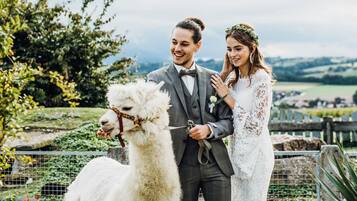Una pareja vestida de novios posa junto a una llama blanca en un entorno al aire libre con follaje y una cerca.