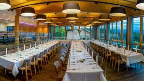 Una larga mesa de comedor dispuesta para un evento formal, con numerosas sillas, amplias ventanas que ofrecen una vista panorámica y un techo de madera con luces colgantes.
