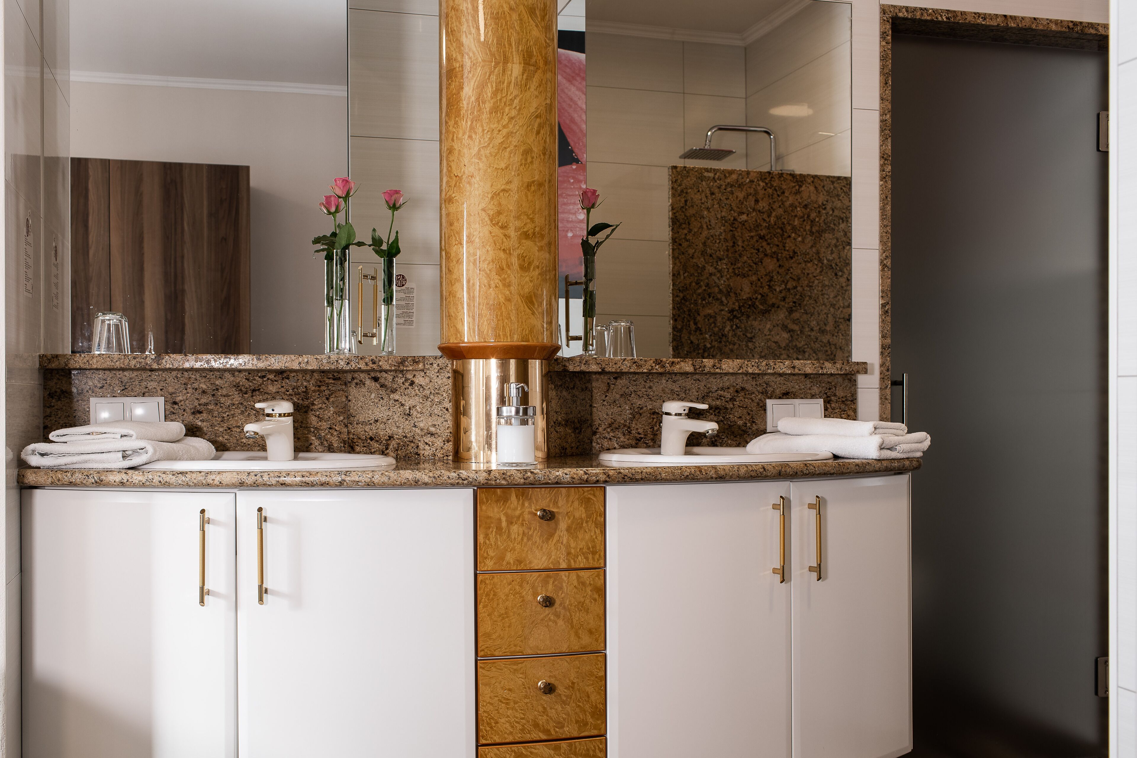A modern bathroom with a granite countertop, two sinks, a large mirror, and a wooden cabinet with gold handles.