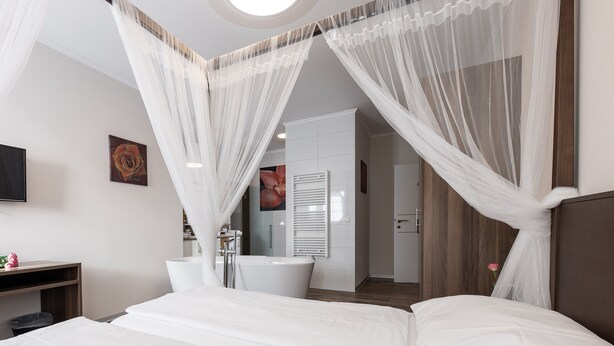 Un dormitorio moderno con una cama grande, una mesita de noche, un televisor y un baño visible a través de una puerta de cristal.