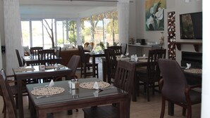 A dining area with multiple tables and chairs, large windows with curtains, and artwork on the walls.