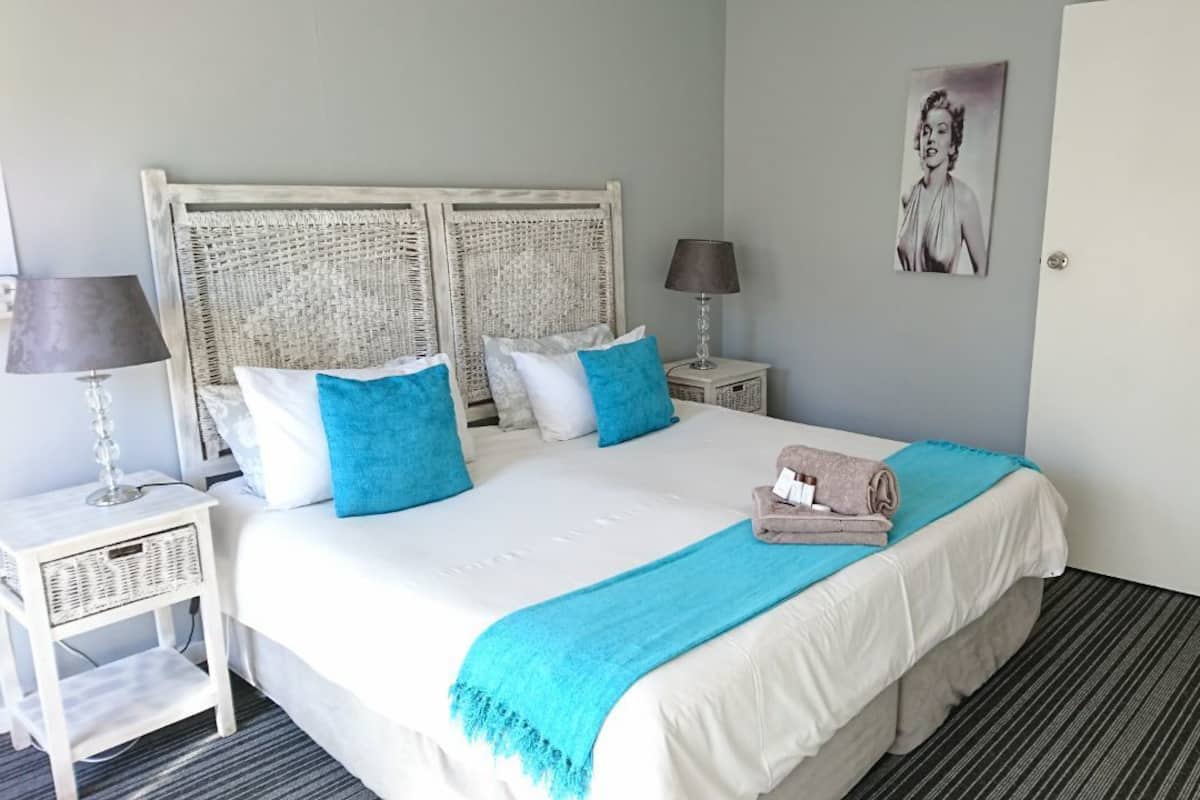 Une chambre d’hôtel bien rangée, avec un grand lit, deux tables de chevet, une lampe, un tableau encadré au mur et une porte.
