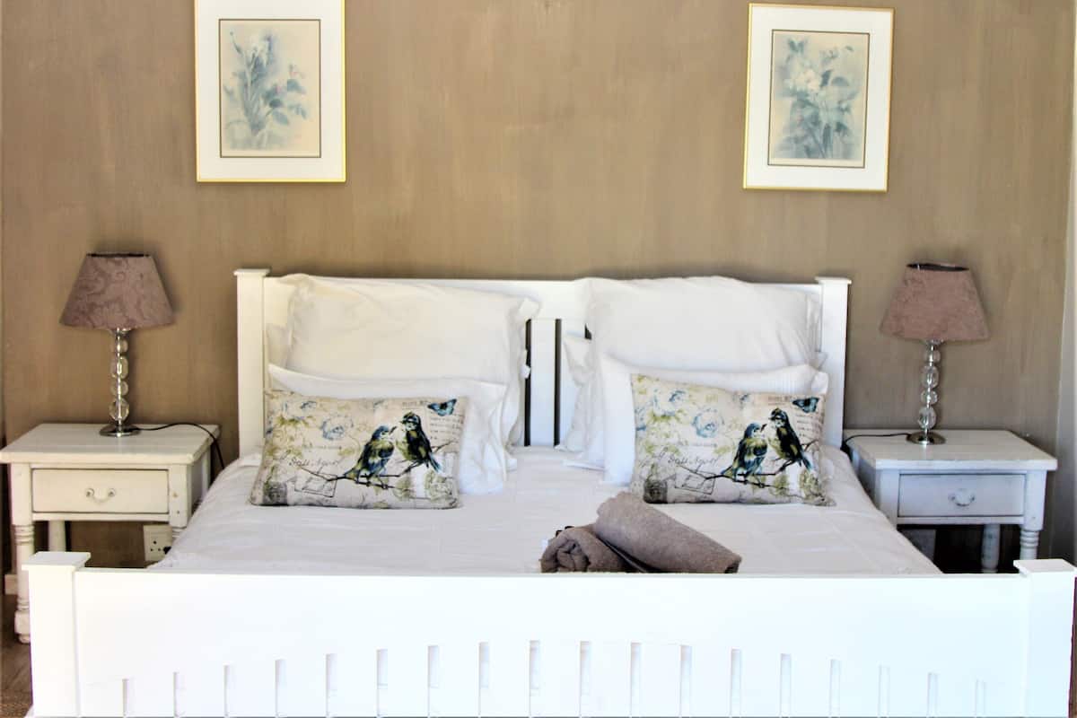 Un lit bien fait, avec du linge de lit blanc et deux coussins décoratifs ornés de motifs d’oiseaux. De chaque côté du lit, il y a une table de chevet avec une lampe, et un cadre photo est accroché au mur, au-dessus du lit.