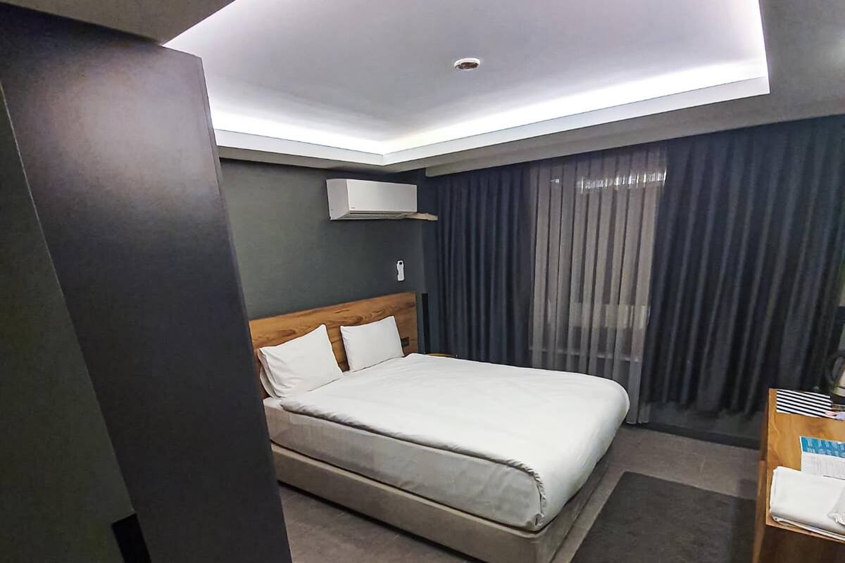 Una habitación de hotel moderna con una cama grande, cabecera de madera, una mesita de noche pequeña, un escritorio con silla y una ventana con cortinas.