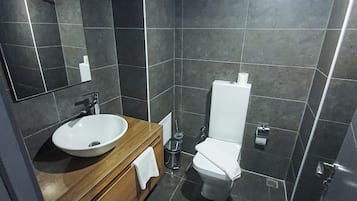 Un baño moderno con baldosas oscuras, un mueble de tocador de madera, un lavabo blanco, un inodoro y una zona de ducha.