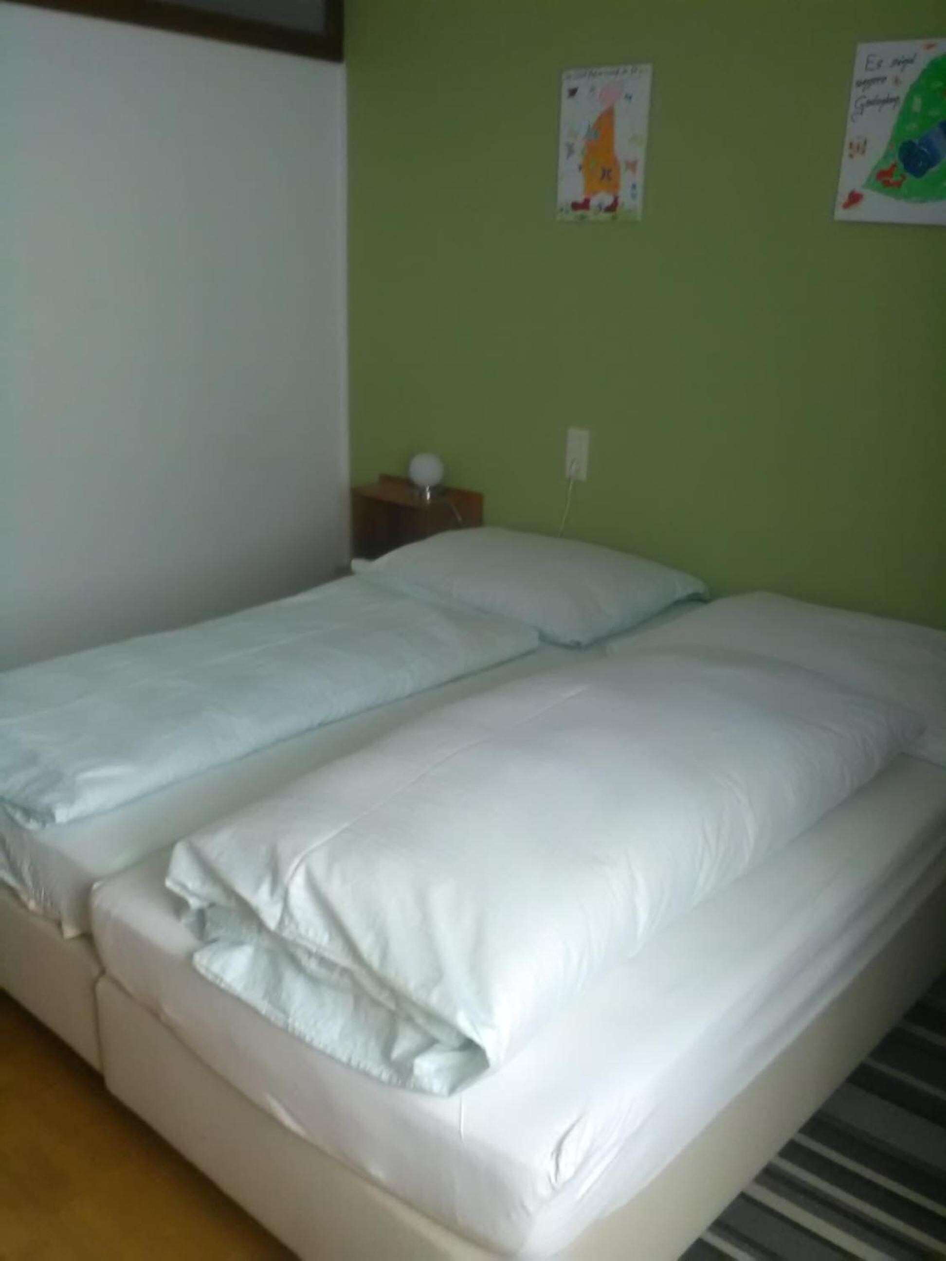 A single-bed room with a green wall, a small bedside table with a lamp, and two posters on the wall.