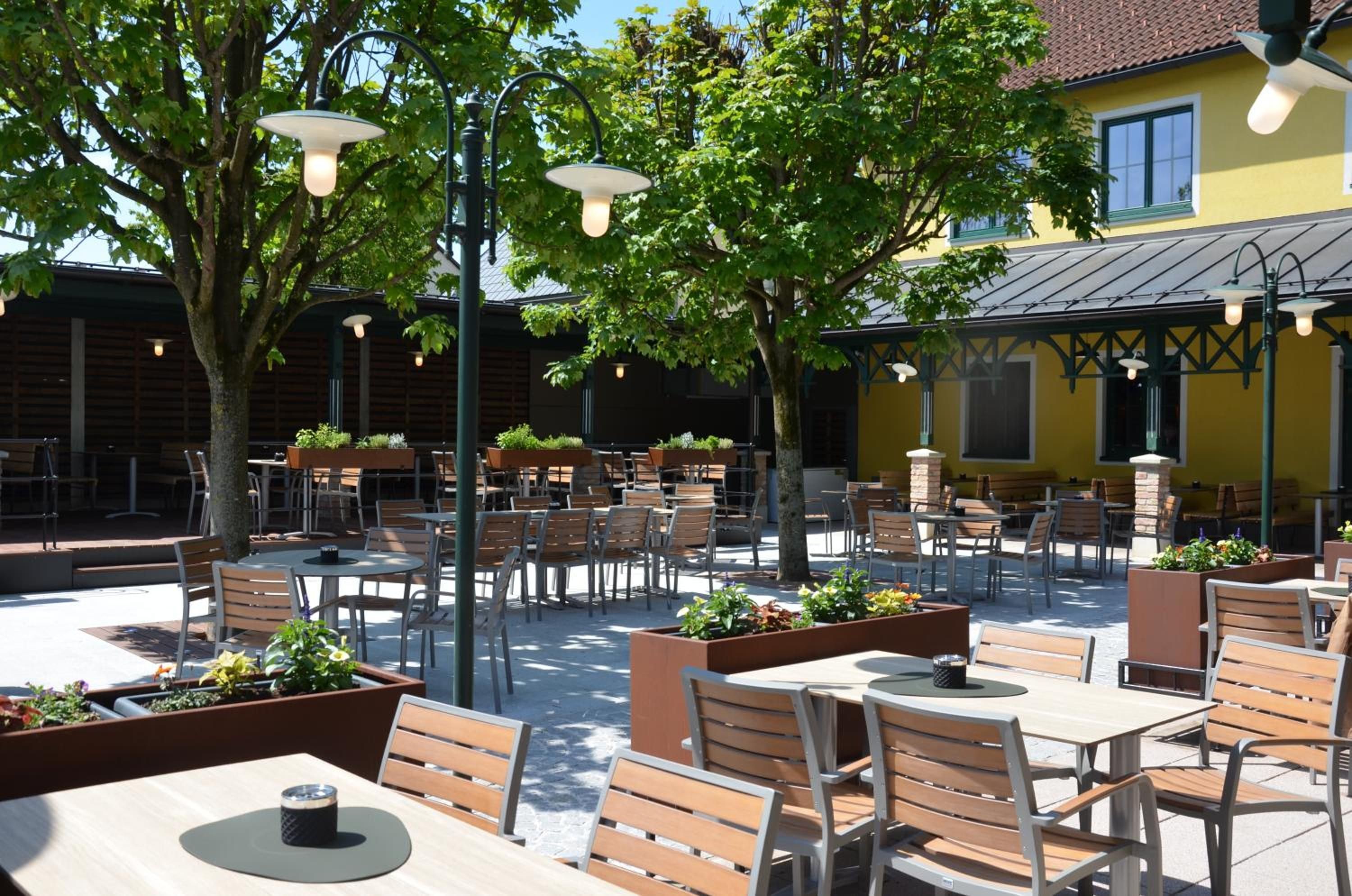 An outdoor seating area with tables and chairs, surrounded by trees and a building with a yellow facade.