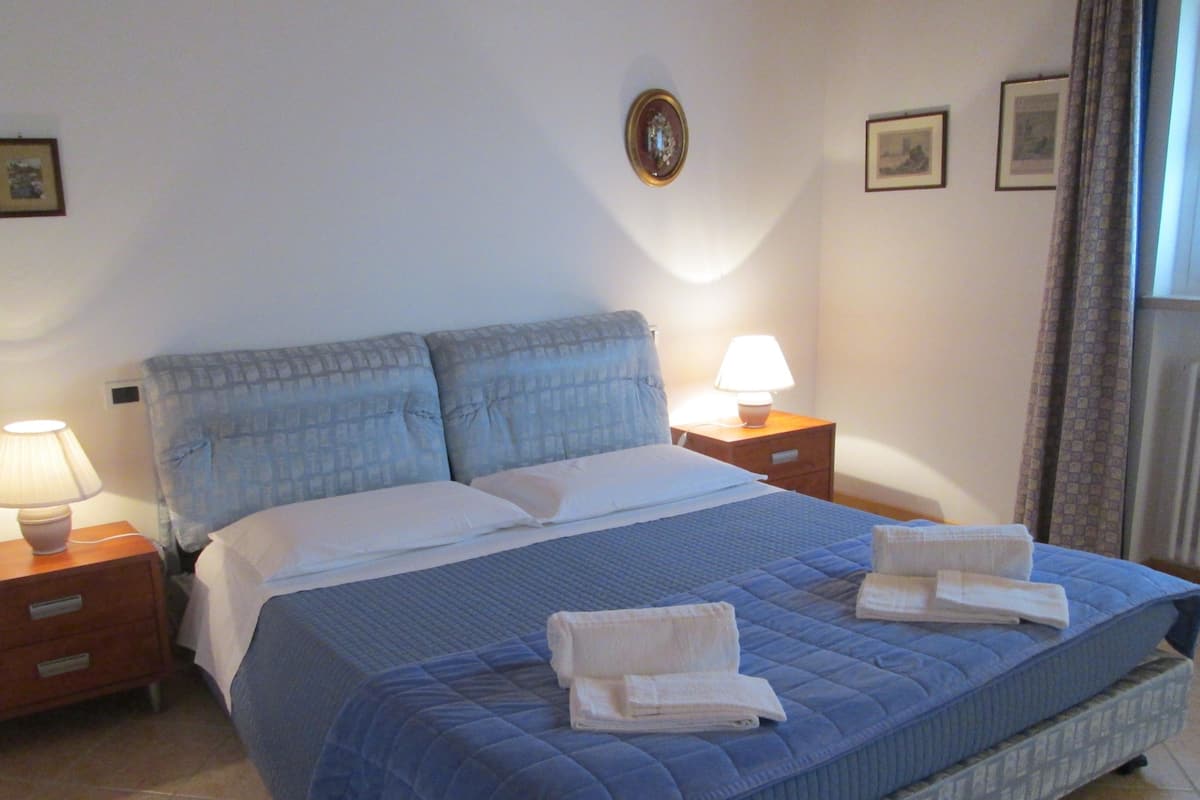 Un lit bien fait, recouvert d’une couette bleue, avec deux oreillers et deux serviettes pliées posées sur le lit. Il y a deux tables de chevet avec des lampes et un petit tableau encadré accroché au mur.