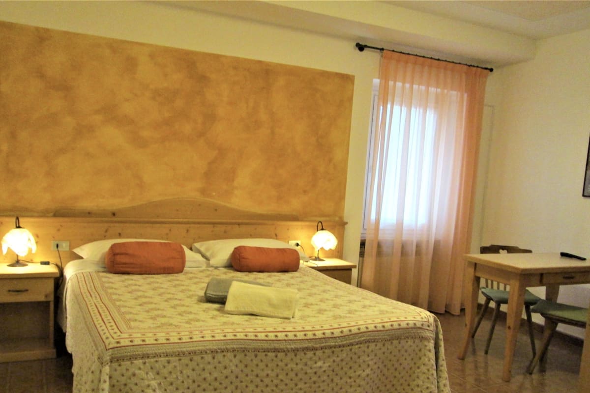 Une chambre d’hôtel avec un grand lit, deux tables de chevet avec des lampes, une tête de lit en bois, un bureau avec une chaise et une fenêtre avec des rideaux.