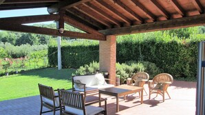 An outdoor patio with wooden furniture, a brick column, and a view of a well-maintained garden.