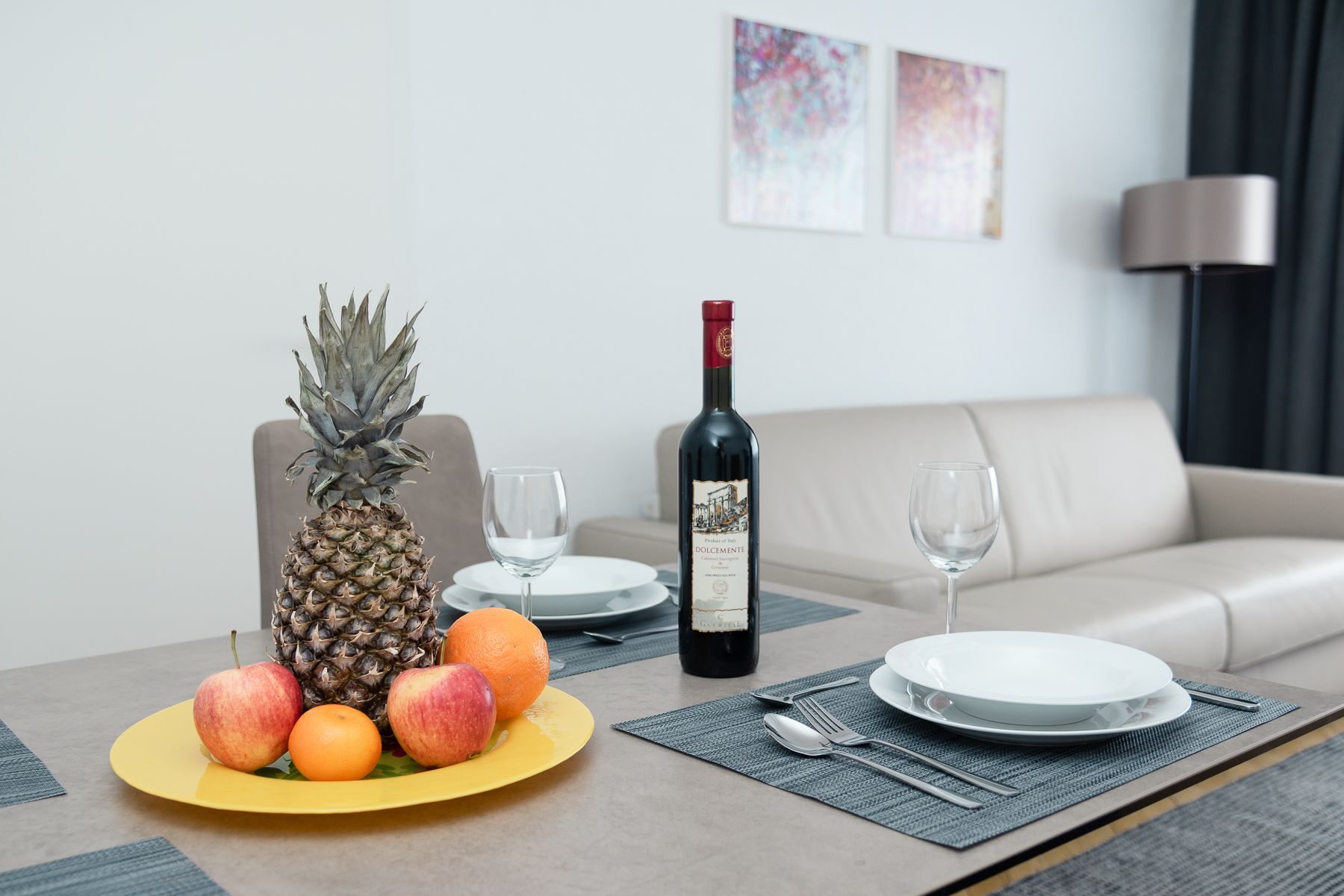 Una mesa de comedor con un plato amarillo de fruta, una botella de vino y una copa.