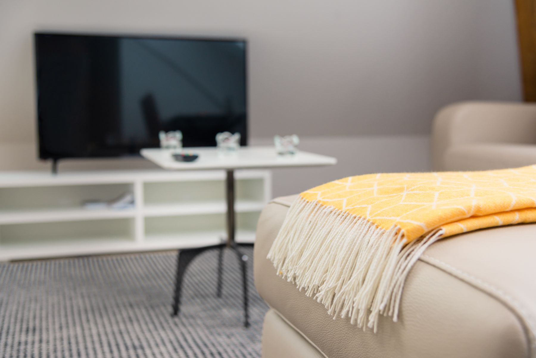 Een moderne woonkamer met een beige bank, een gele plaid, een flatscreen-televisie en een witte bijzettafel met een glazen blad.