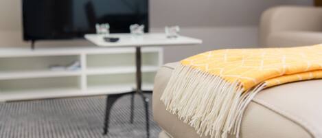 Una sala moderna con un sofá color beige, una manta amarilla, un televisor de pantalla plana y una mesita auxiliar blanca con cubierta de vidrio.