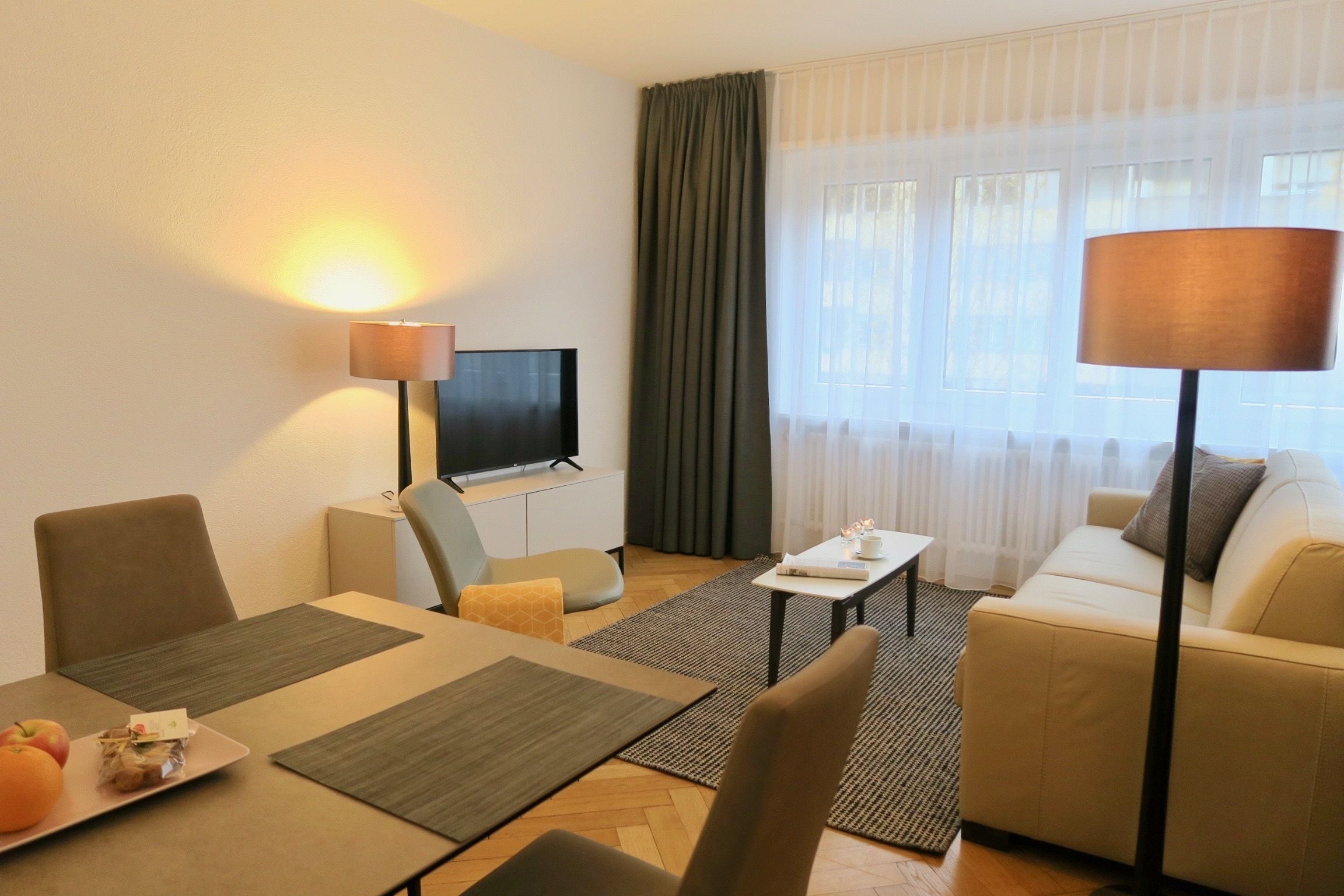 Ein modernes Hotelzimmer mit einem Flachbildfernseher, einer Couch, einem Esstisch mit Platzsets und einer Lampe.