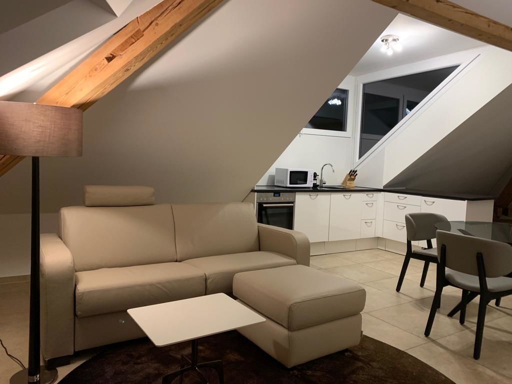 Un espacio habitable estilo loft con sofá, reposapiés y una pequeña mesa. Se ve un comedor con sillas, y una cocina con armarios blancos y una ventana al fondo.