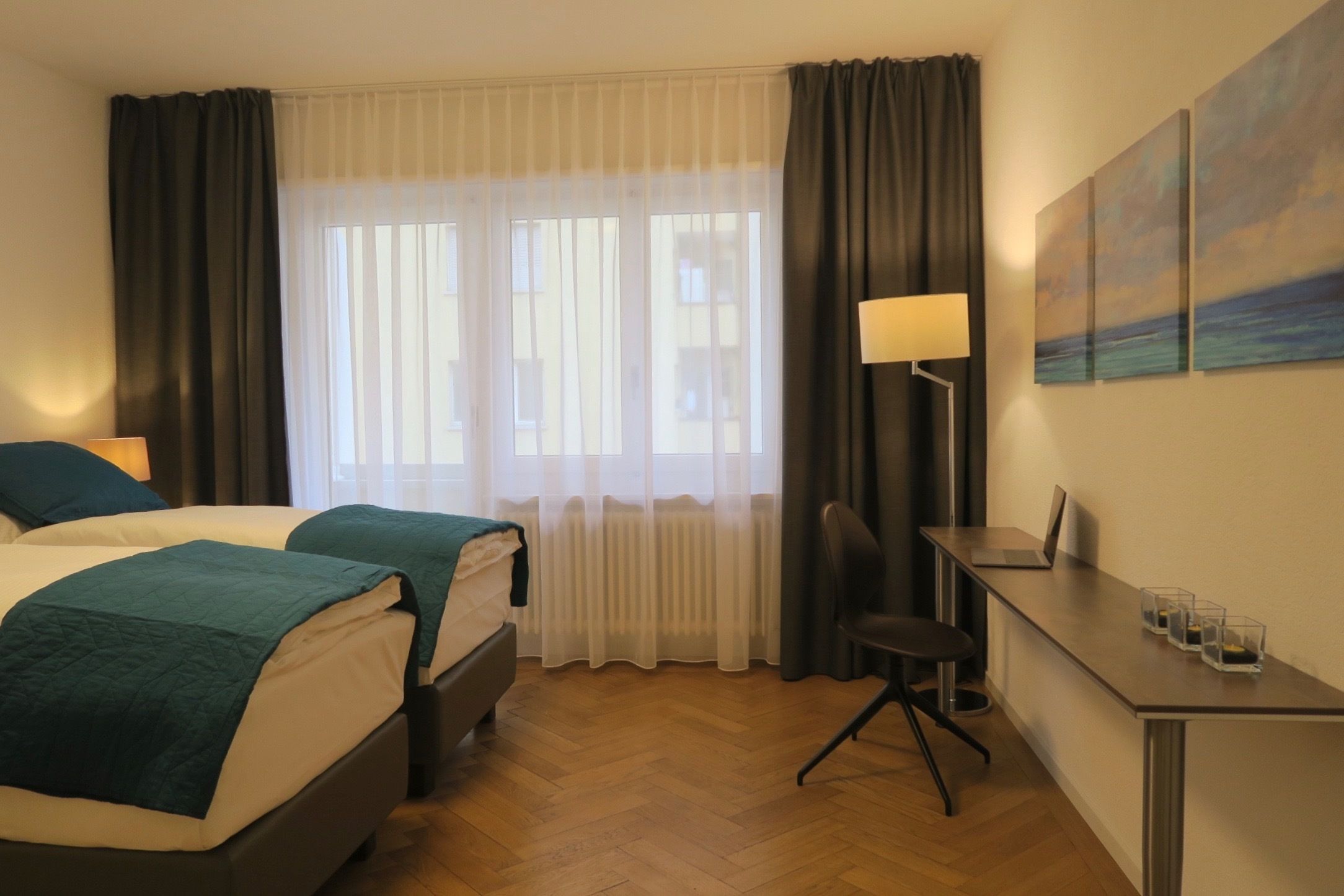 A hotel room with two beds, a desk, a chair, a lamp, and a painting on the wall.