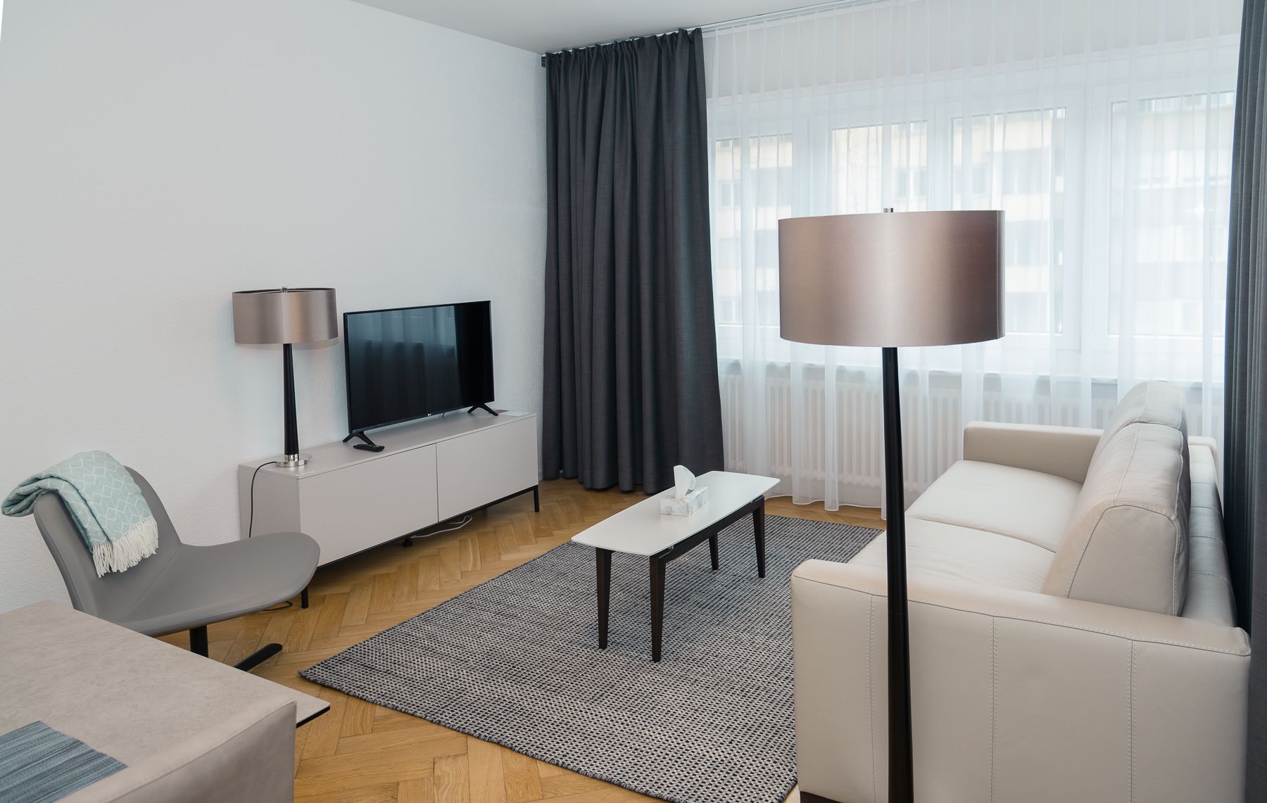 A modern living room with a flat-screen TV, a sofa, a coffee table, a lamp, and a large window with curtains.