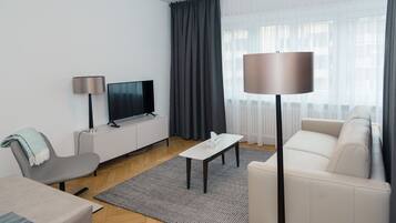 A modern living room with a flat-screen TV, a sofa, a coffee table, a lamp, and a large window with curtains.