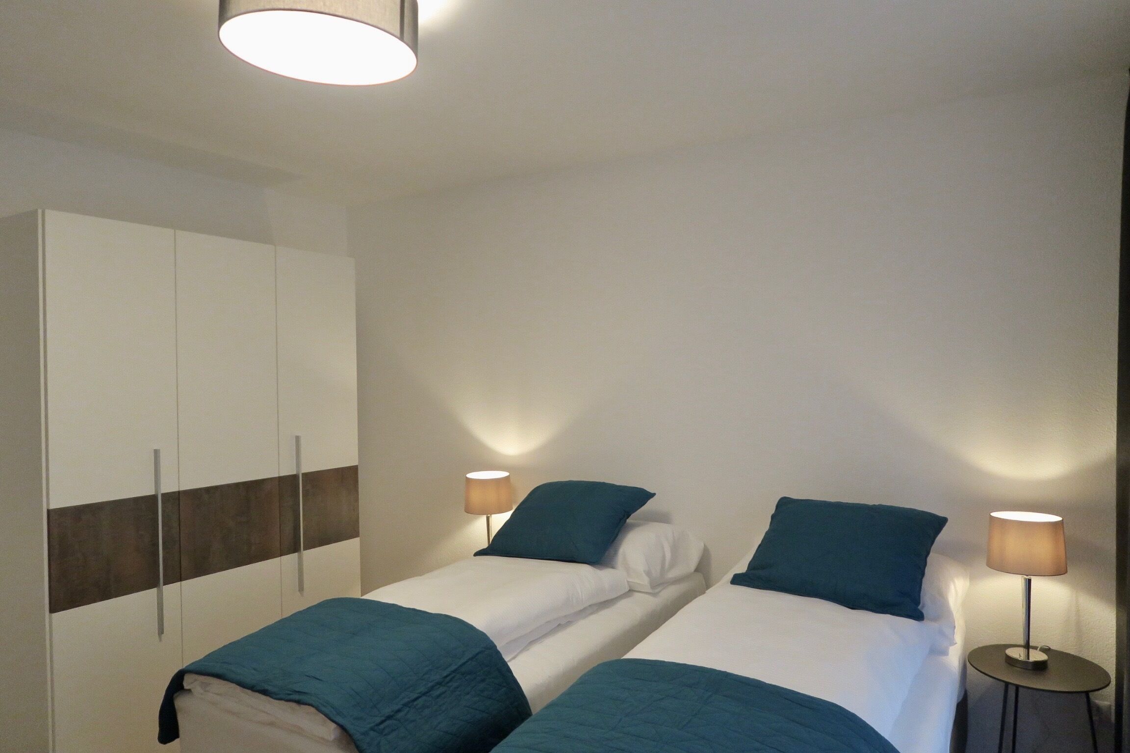A room with two beds, each with a blue blanket and pillow, a wardrobe, and two bedside lamps.