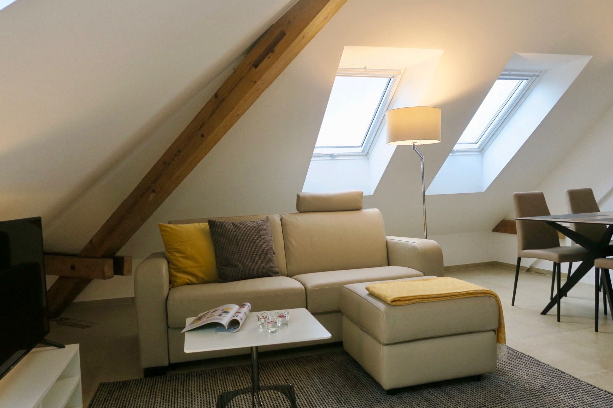 Un moderno espacio tipo loft con sofá, puf y mesa de comedor. Cuenta con tragaluces y una lámpara de pie.