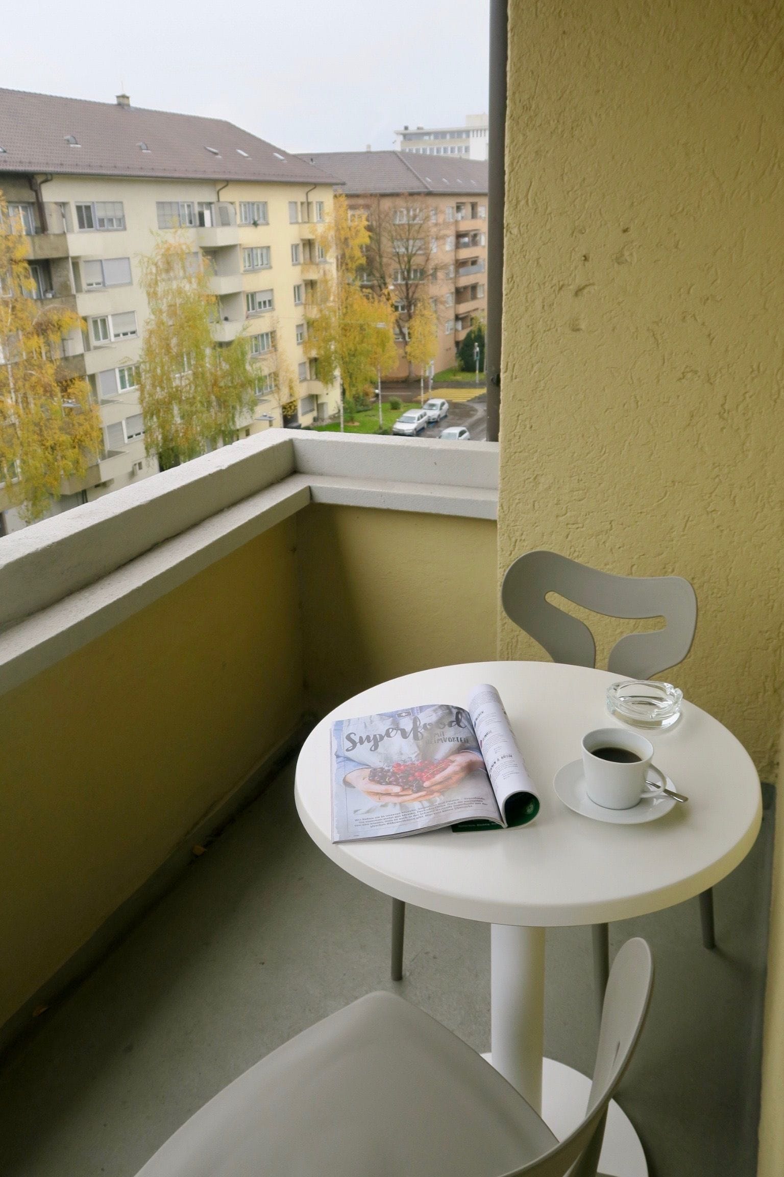 Uma varanda com uma mesa e cadeira brancas, uma revista, uma xícara de café e vista para prédios de apartamentos e árvores.