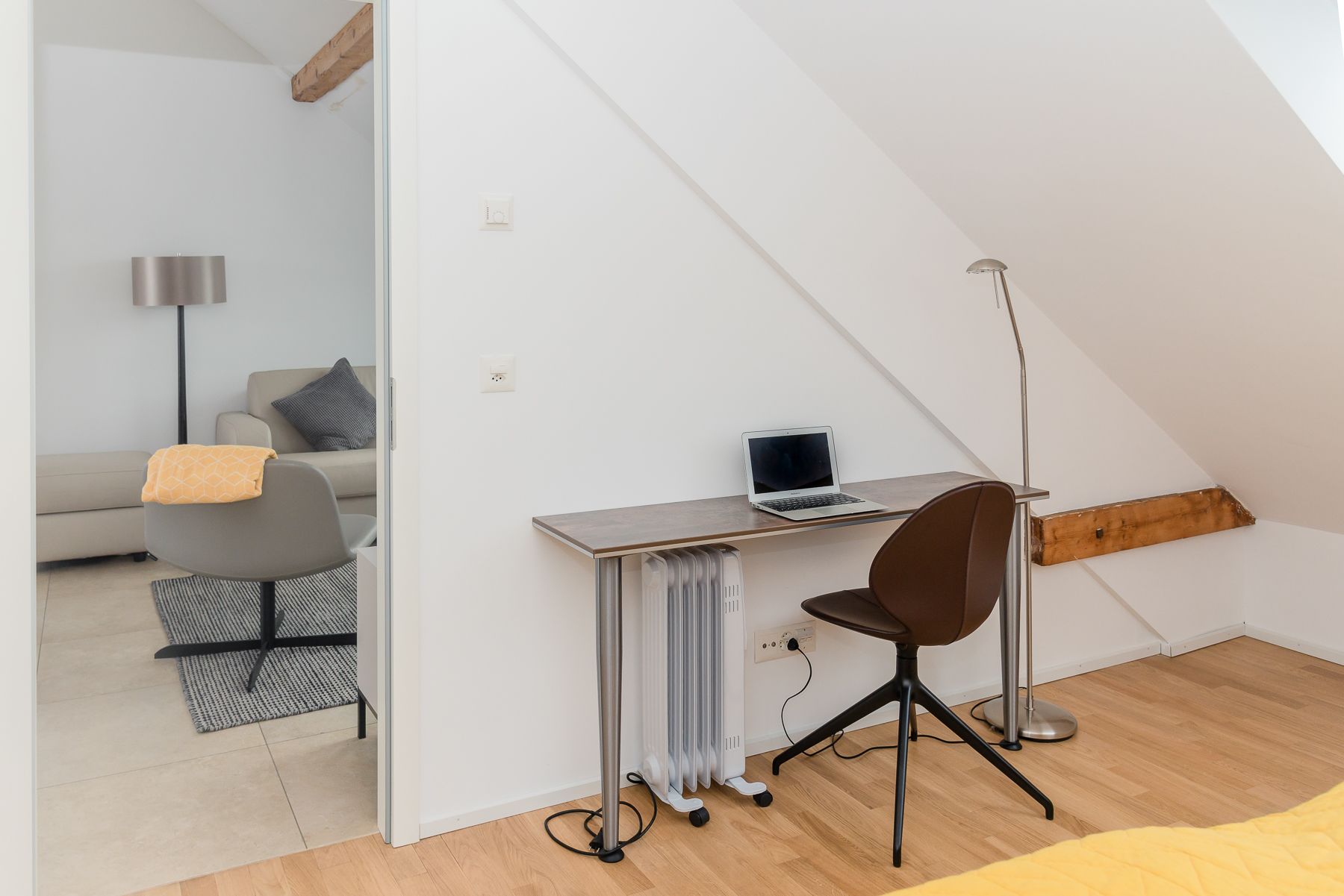 Une chambre de style loft moderne, avec un plafond en pente, un bureau intégré doté d’un ordinateur portable, un fauteuil de bureau marron et un radiateur.