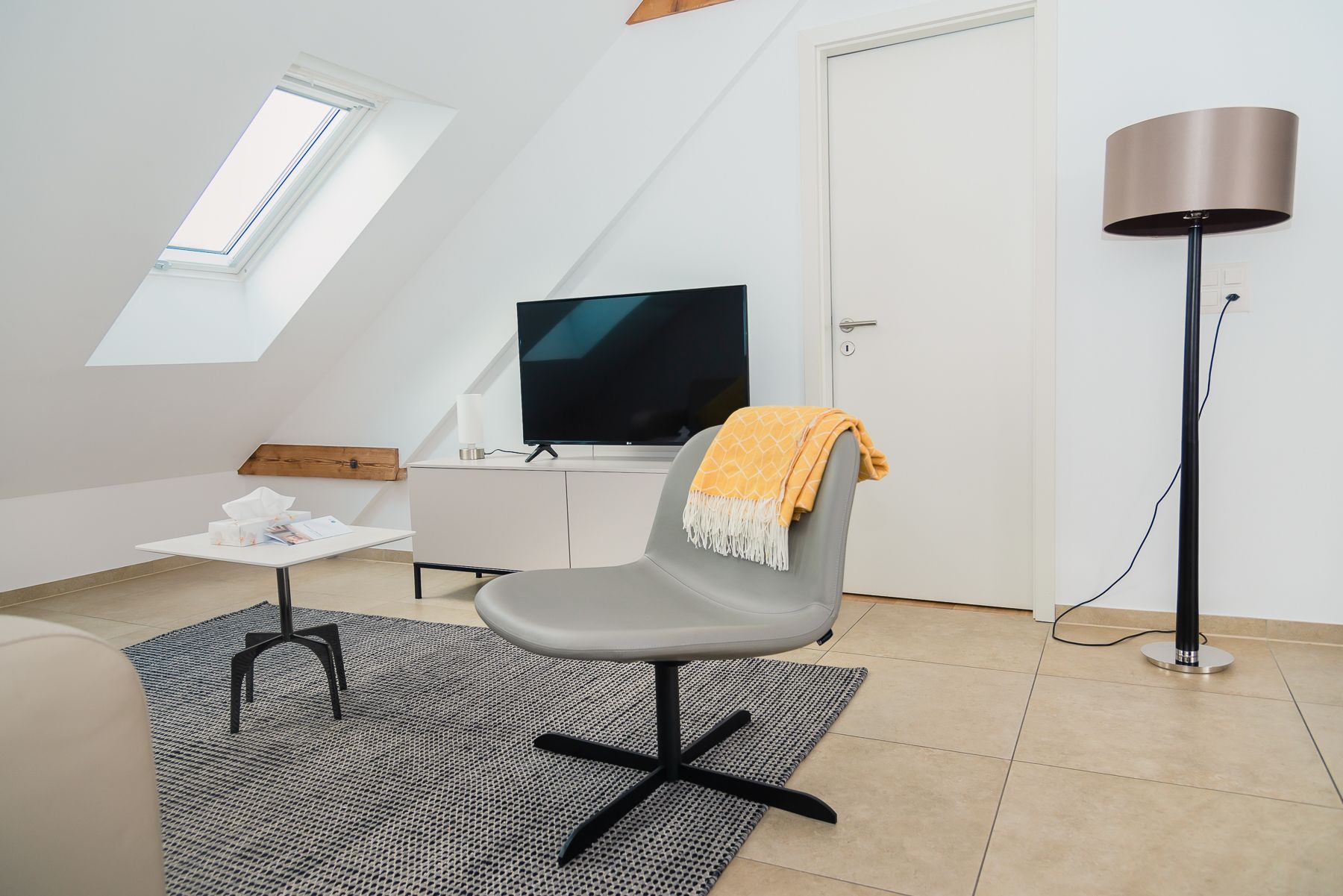 A modern, well-lit room with a sloped ceiling, a flat-screen TV on a white cabinet, a grey chair with a yellow throw, a small table with a tissue box, and a floor lamp.