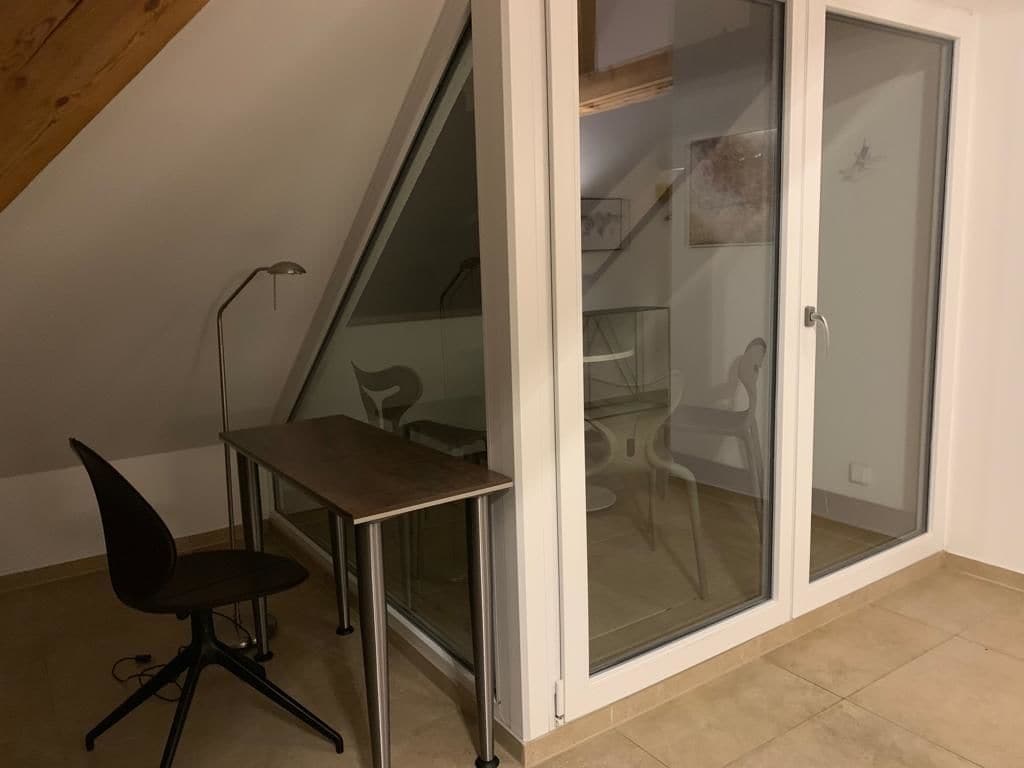 A room with a sloped ceiling, a desk with a chair, and a sliding glass door leading to a balcony with chairs.