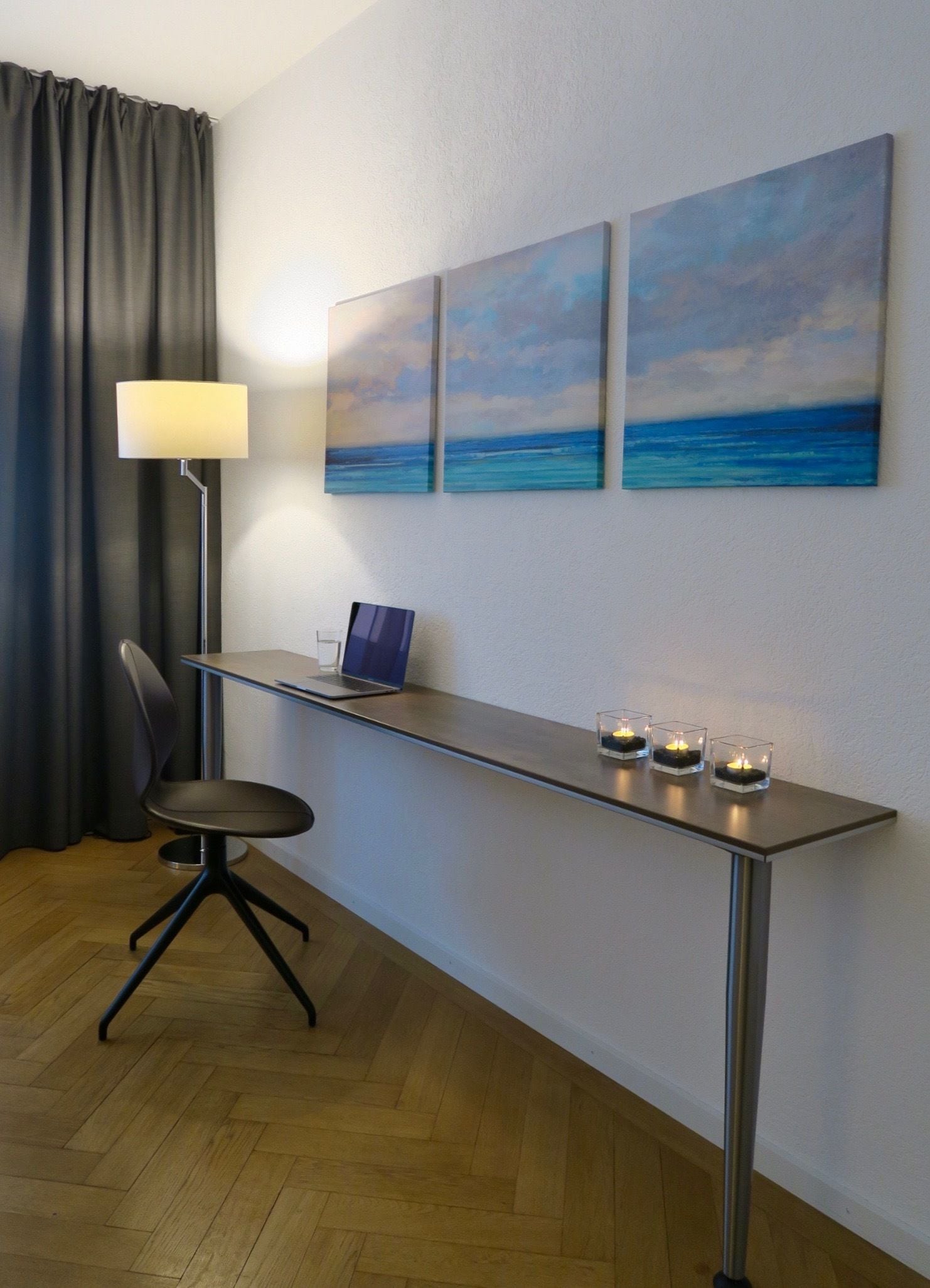 A modern room with a desk, a laptop, and three paintings of the ocean.
