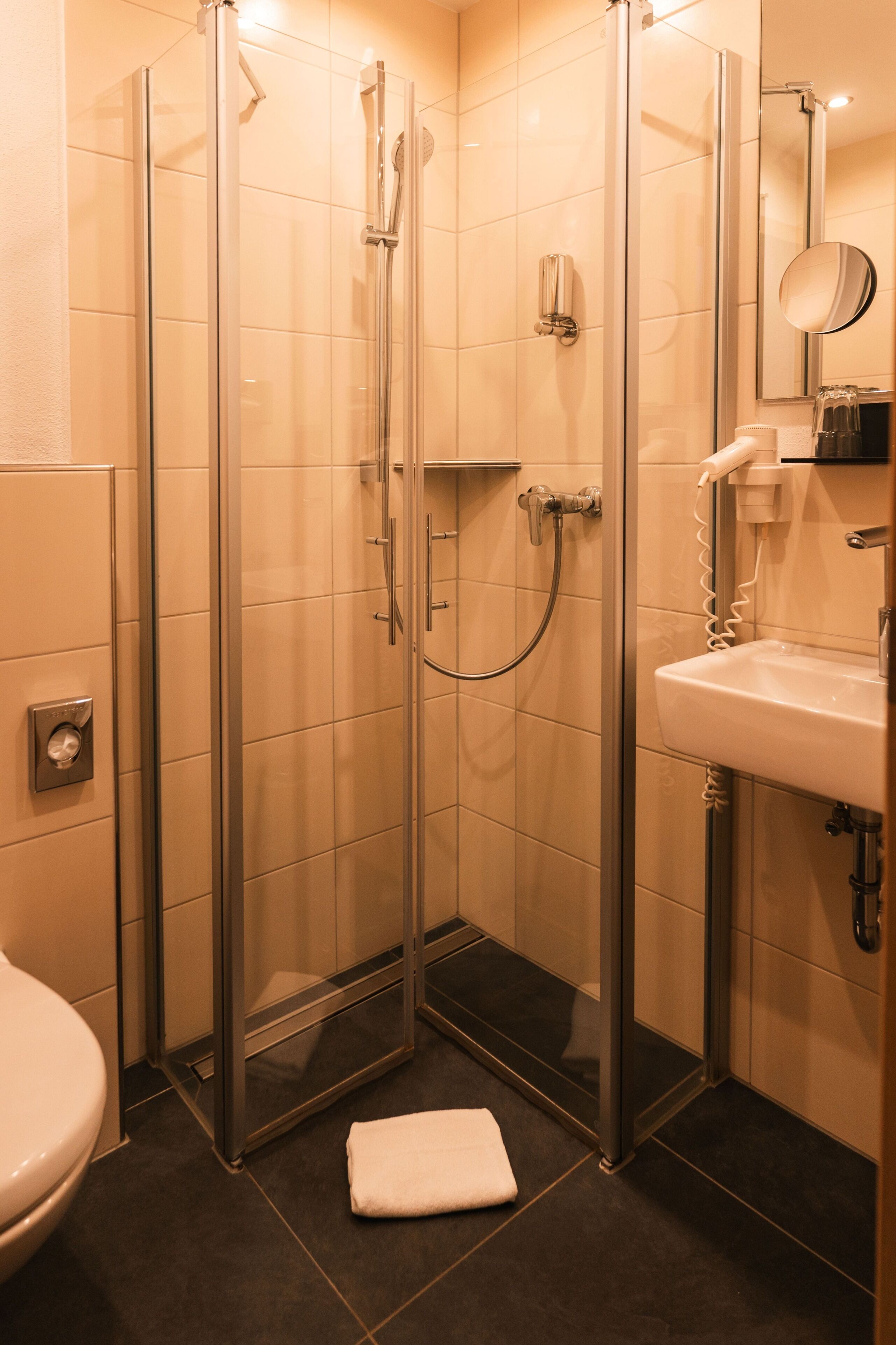A glass-enclosed shower stall with a white towel on the floor, a sink with a mirror, and a toilet.