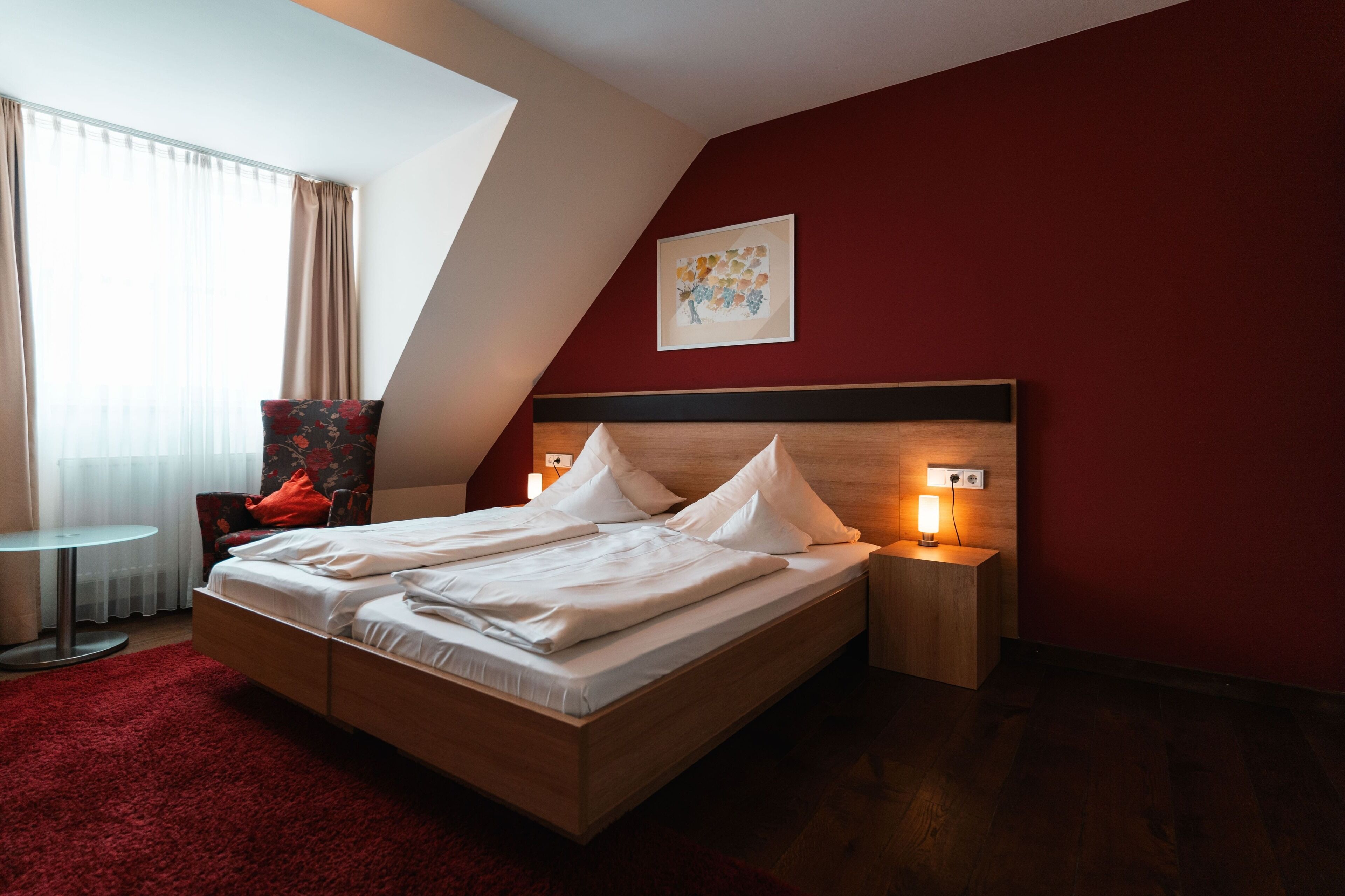 Una habitación de hotel moderna con una cama de madera, una alfombra roja, una mesita redonda y un sillón rojo.