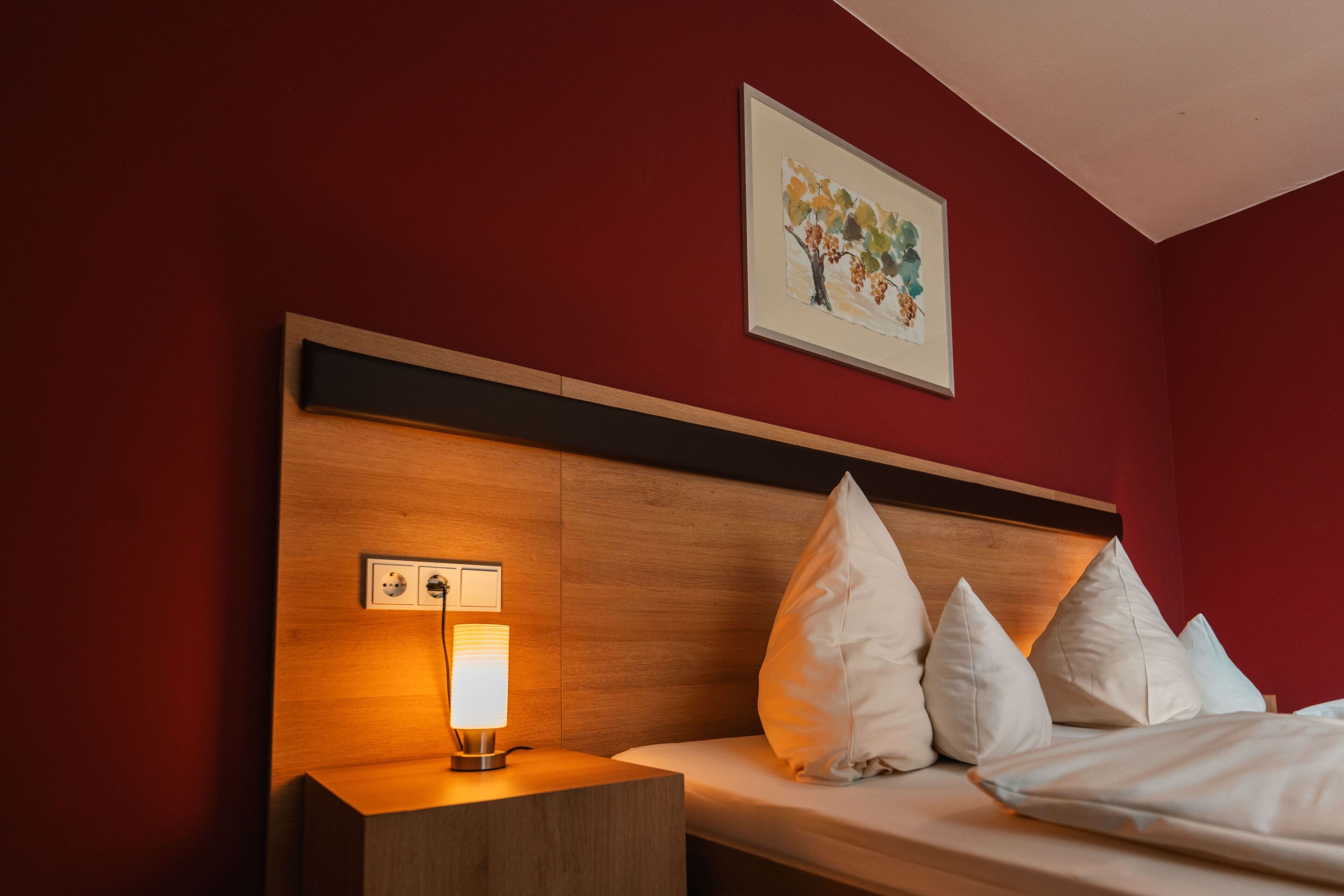 Quarto de hotel com cabeceira de madeira, mesa de cabeceira com abajur e cama com almofadas brancas.