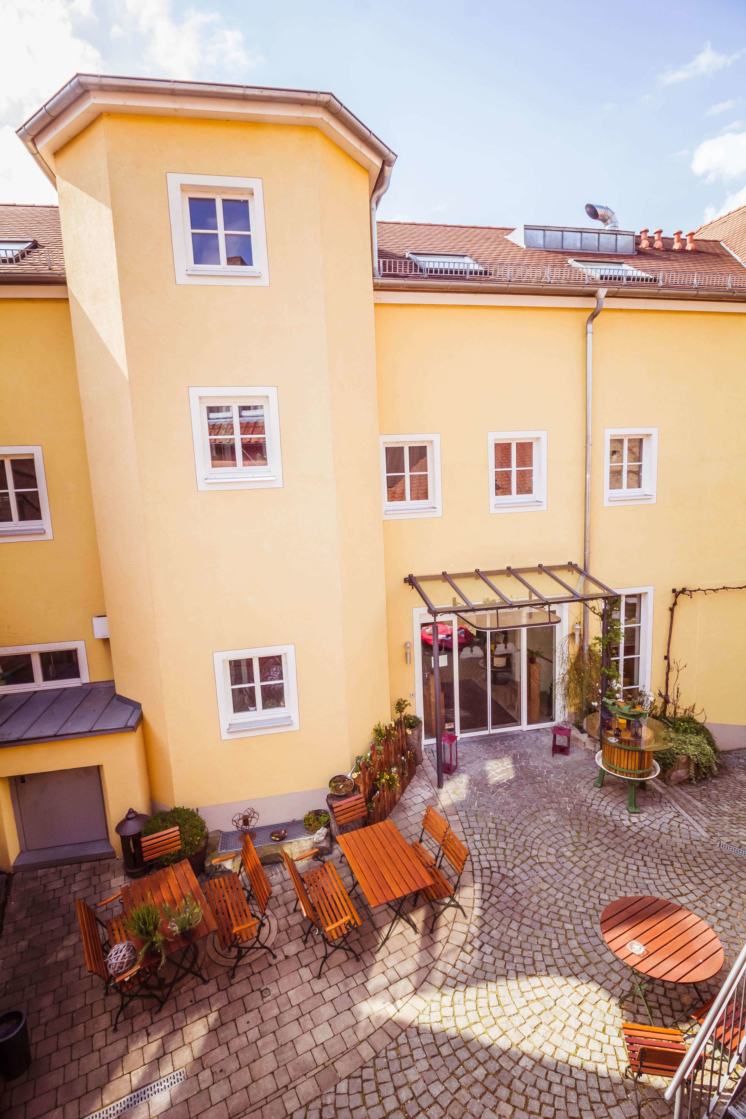 A yellow building with multiple windows, a covered patio area with wooden furniture, and a small garden.