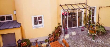 A yellow building with multiple windows, a covered patio area with wooden furniture, and a small garden.