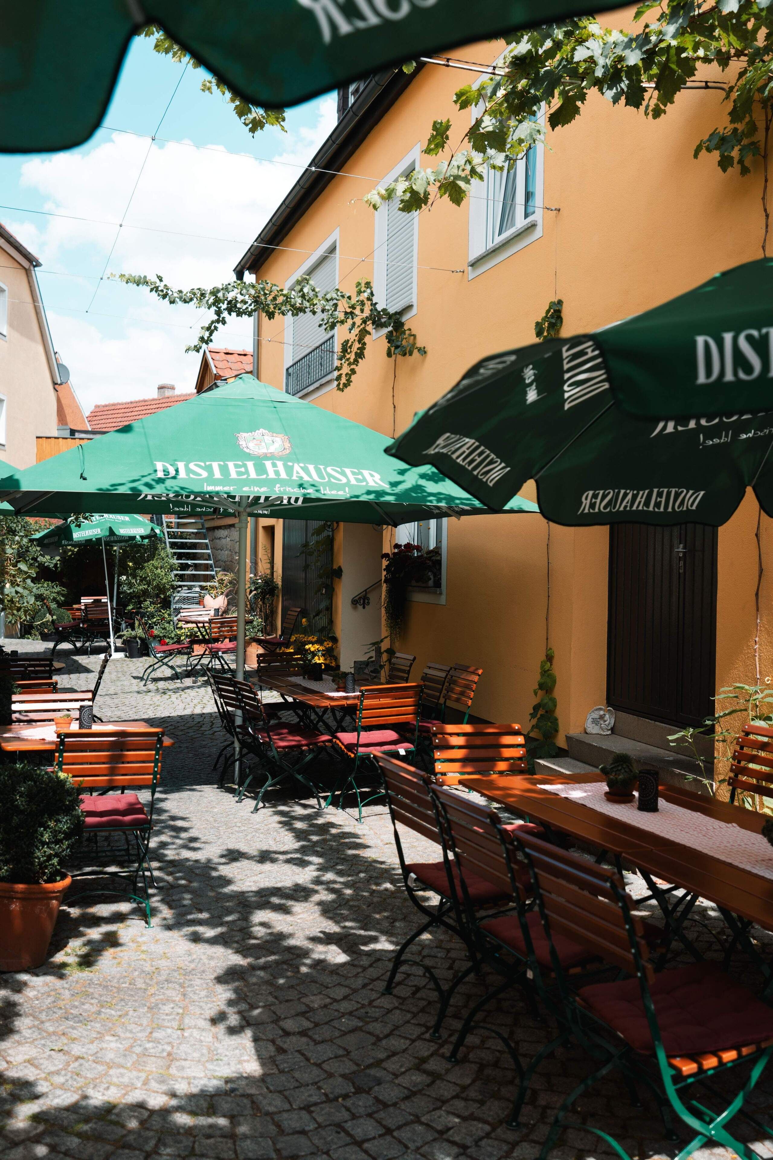 Ein Straßencafé mit Holztischen und -stühlen, grünen Sonnenschirmen und einem Kopfsteinpflasterweg.