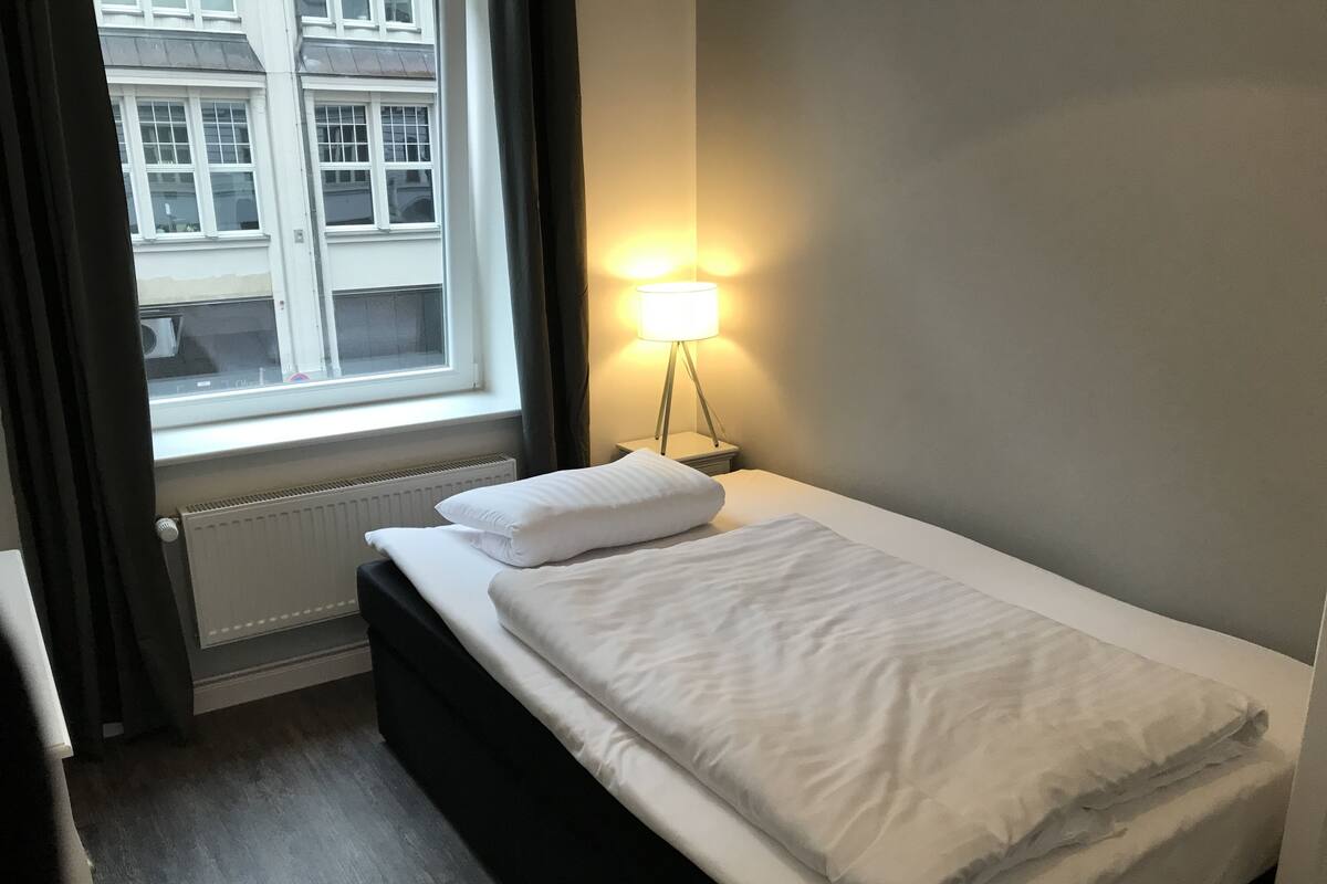 Una cama bien hecha con sábanas blancas, una lámpara de noche, una ventana con cortinas y un radiador.