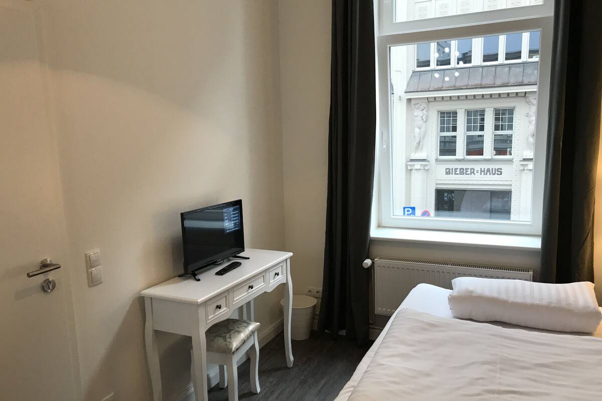 Habitación de hotel con una cama, un escritorio con televisión, una ventana con vista a un edificio y una puerta.