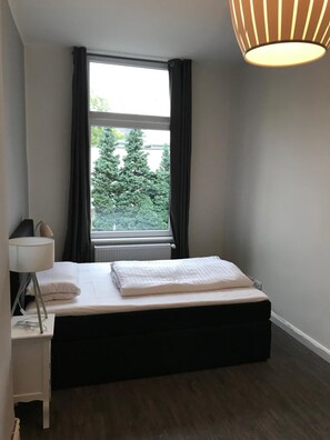 A bedroom with a bed, a bedside table, a lamp, a window with curtains, and a view of trees outside. - Hotel Fame am Hauptbahnhof (Hamburg)