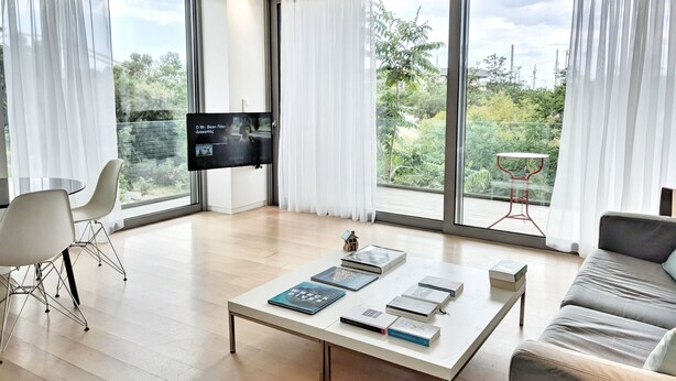 Un salon moderne doté de grandes fenêtres, d’un téléviseur à écran plat, d’un canapé, d’une table basse avec des livres et d’un coin repas avec des chaises.