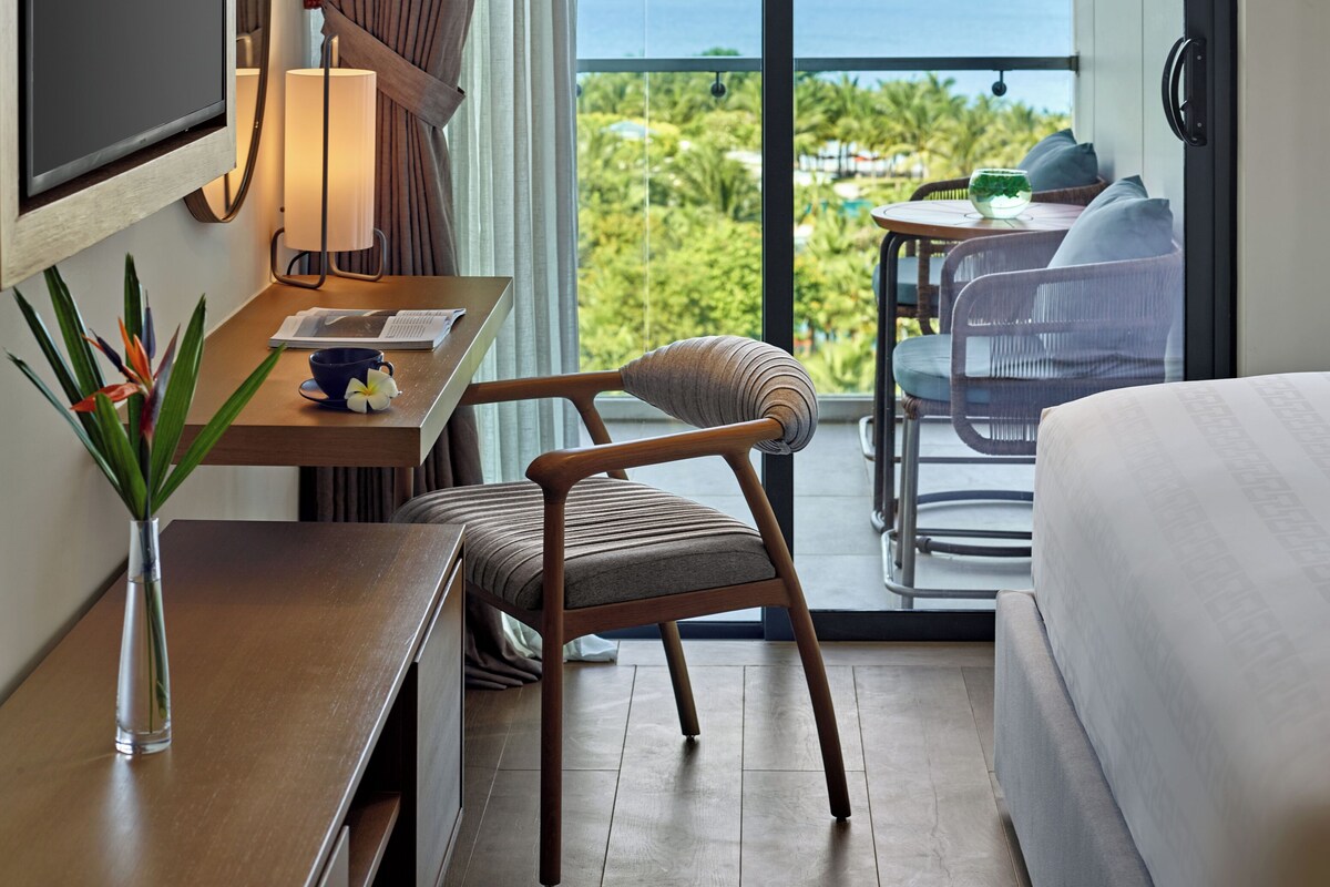 Habitación de hotel con cama, escritorio con televisor, silla, mesa con vaso y balcón con vista al océano.