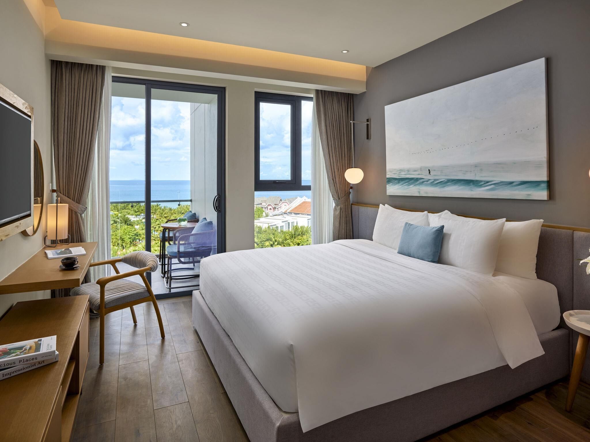 A hotel room with a large bed, a desk with a chair, a TV, and a view of the ocean.