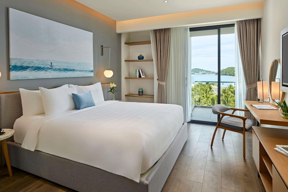 Habitación de hotel moderna con una cama grande, un escritorio con silla, una estantería y un ventanal con vistas al mar.