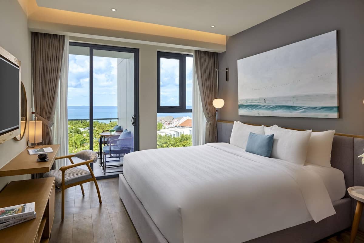 Habitación de hotel con una cama grande, un escritorio con silla, televisión y balcón con vista al océano.