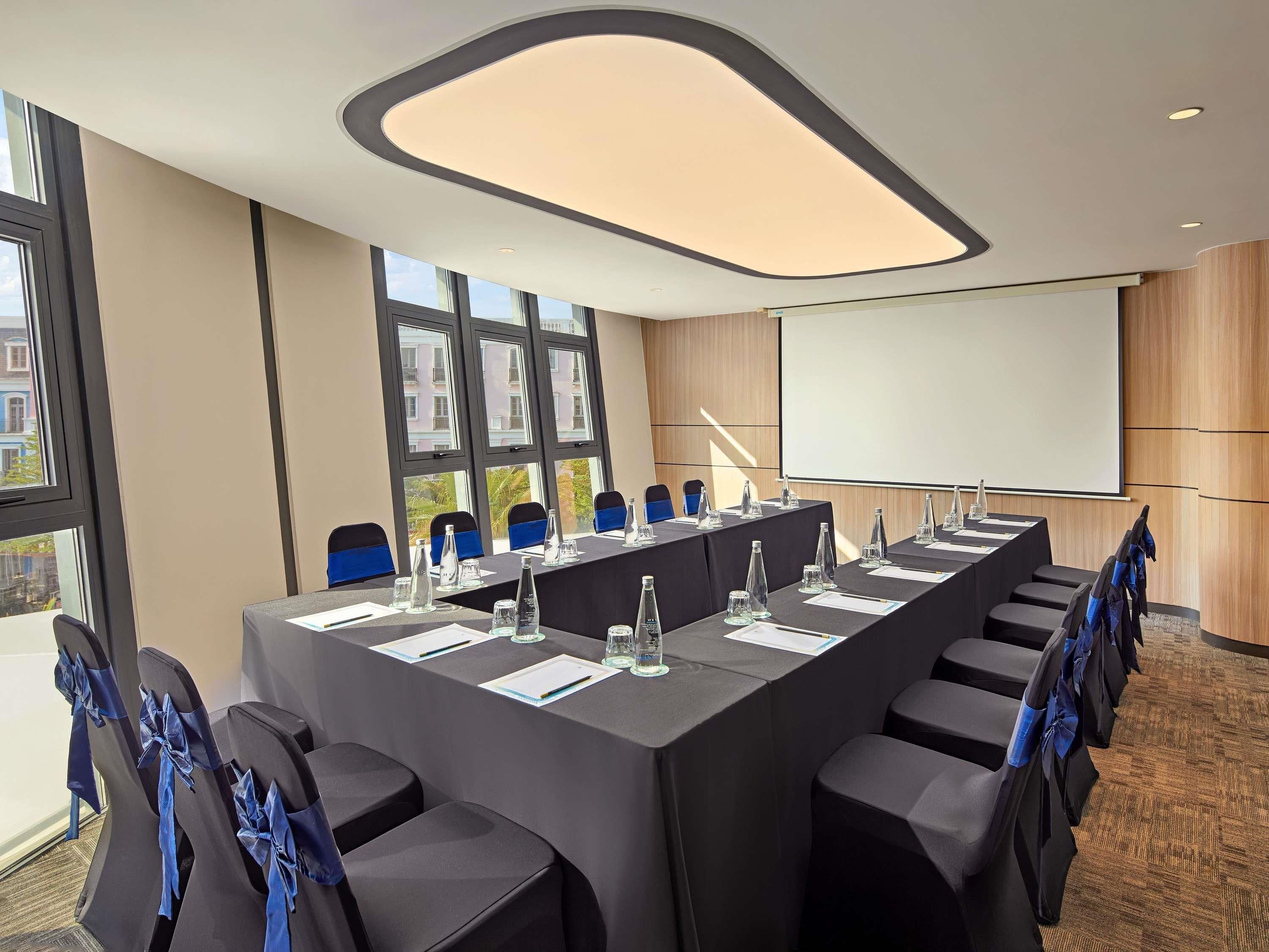 Un salón de conferencias con una larga mesa, sillas, una pantalla de proyector y un amplio ventanal.