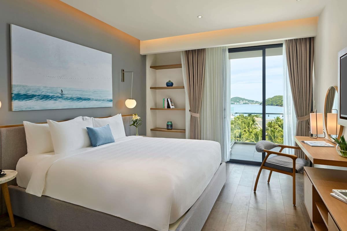 Habitación de hotel moderna con una cama grande, un escritorio con silla, una estantería y un ventanal con vistas al mar.
